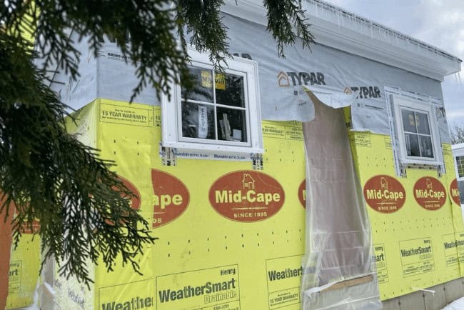 Construction site featuring a house under renovation with exposed windows, yellow Mid-Cape building materials, and Typar weather barrier, illustrating Coast Carpentry's home improvement services in Cape Cod, MA.
