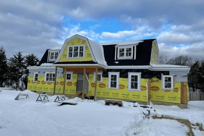 Newly constructed house with yellow insulation wrap, snow-covered ground, and cloudy sky, showcasing Coast Carpentry's home improvement and deck building expertise in Cape Cod, MA.