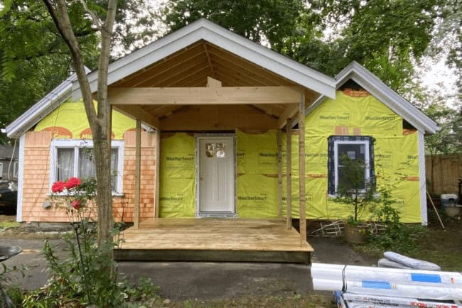 House under construction with a wooden porch, orange siding, and yellow weather barrier, showcasing carpentry work for home improvement projects in Cape Cod.