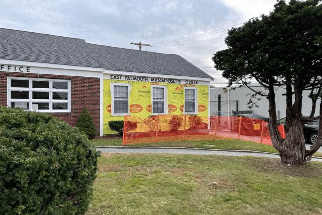Building exterior in East Falmouth, Massachusetts, featuring bright yellow wall with signage, surrounded by orange construction fencing and greenery.