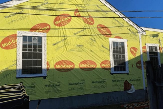 House undergoing renovation with yellow weather-resistant barrier featuring "Mid-Cape" branding, two windows visible, and construction materials in the foreground.