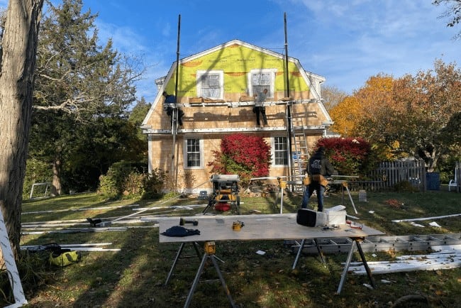 Construction workers performing home improvement on a residential property in Cape Cod, with scaffolding and tools visible, emphasizing deck building and exterior renovations.
