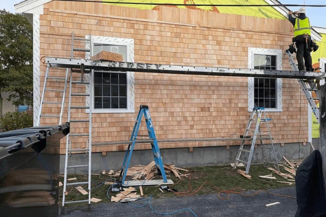 Construction worker installing cedar shingles on a house exterior, scaffolding and ladders visible, with tools and debris on the ground, representing Coast Carpentry's home improvement services.