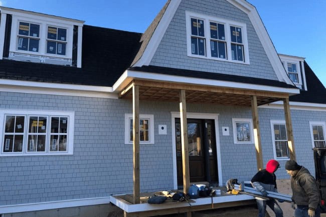 Construction workers building a wooden deck in front of a newly constructed home with gray siding and multiple windows, emphasizing Coast Carpentry's deck building services.