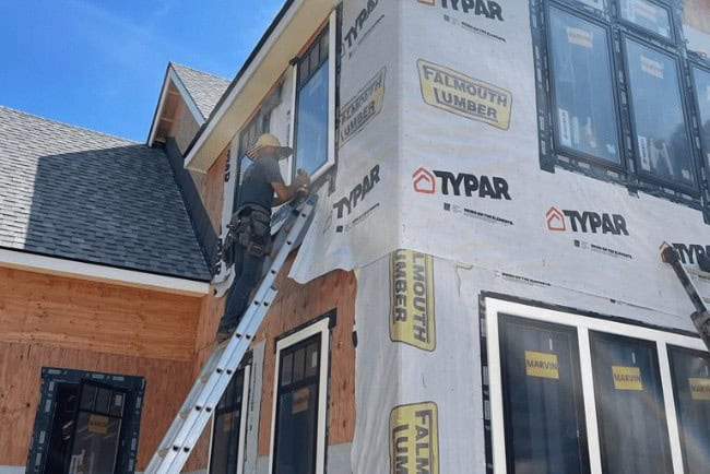 Construction worker installing windows on a residential building, showcasing Typar house wrap and Falmouth Lumber branding, emphasizing Coast Carpentry's home improvement services in Cape Cod, MA.