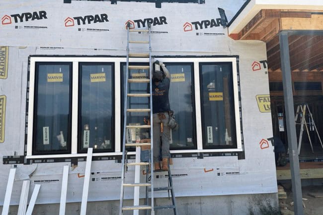 Construction worker installing Marvin windows on a residential building, using a ladder, with Typar house wrap visible.