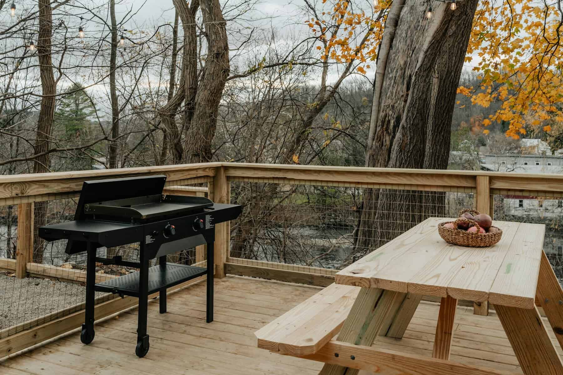 Black outdoor grill on wooden deck with picnic table and basket of apples, surrounded by trees and autumn foliage.
