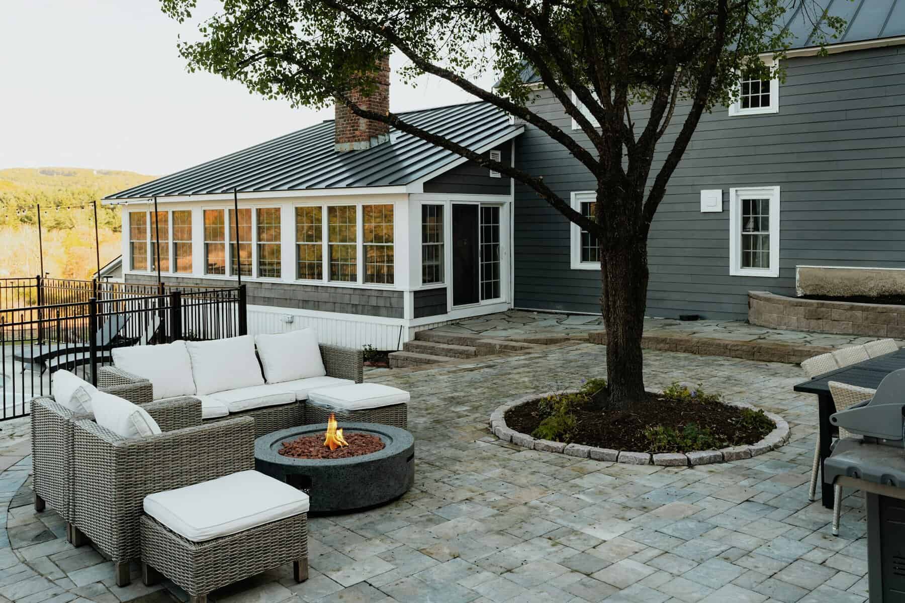 Patio area featuring a fire pit surrounded by modern outdoor seating, with a gray house and lush greenery in the background, illustrating outdoor living space design for home improvement.