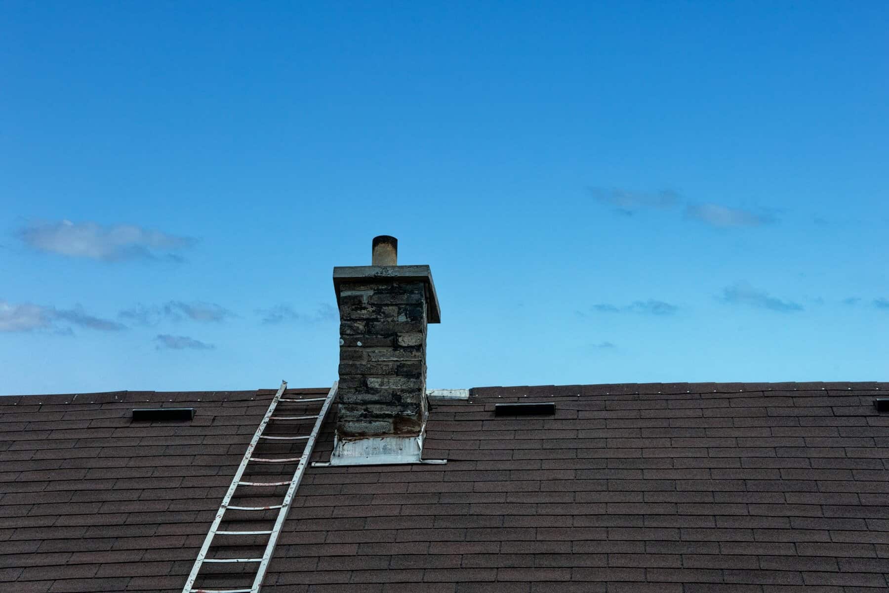 Chimney on a sloped roof with a ladder against the side, under a clear blue sky, illustrating roofing and chimney maintenance.
