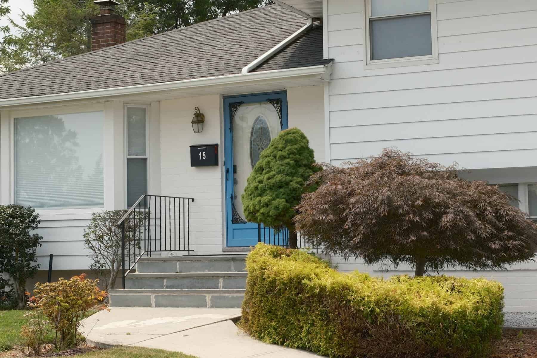 Front entrance of a home featuring a blue door, house number 15, black railing, and landscaped shrubs, illustrating residential construction and curb appeal.