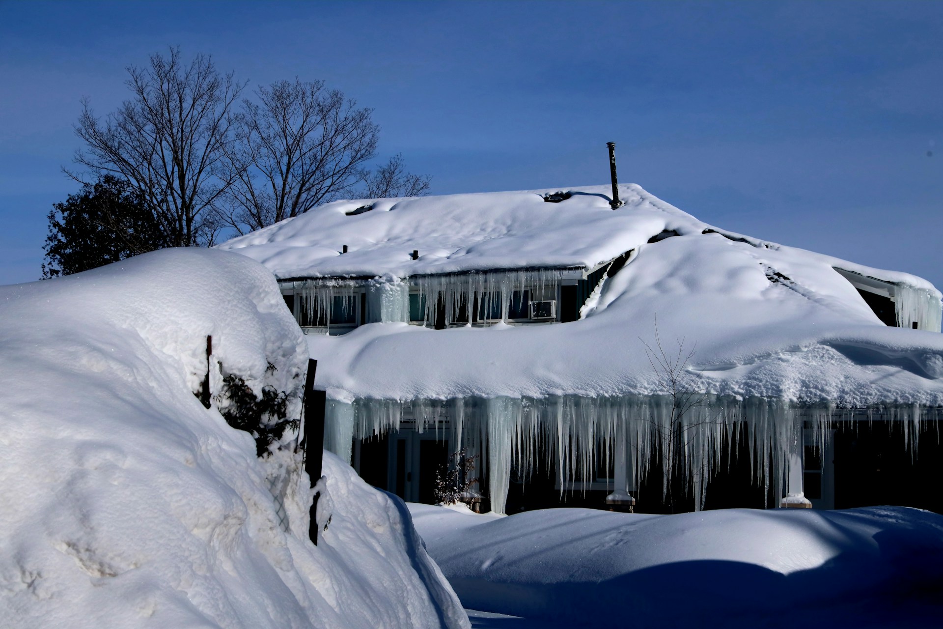 Snow-covered roof with icicles hanging over windows, surrounded by deep snow, illustrating winter weather challenges for home maintenance.
