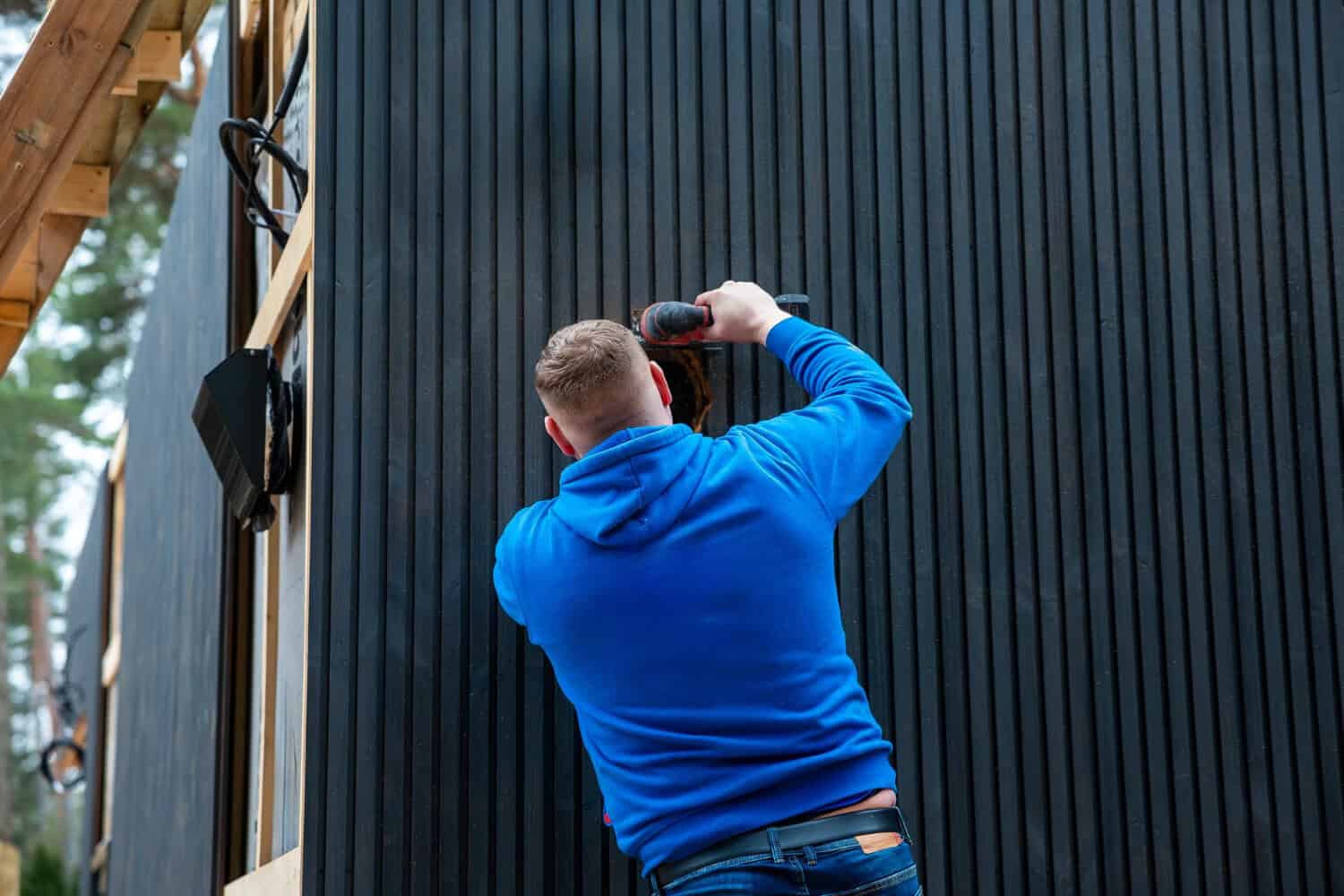 Person in blue hoodie installing siding on a wooden structure, emphasizing home improvement and construction techniques relevant to Cape Cod homes.