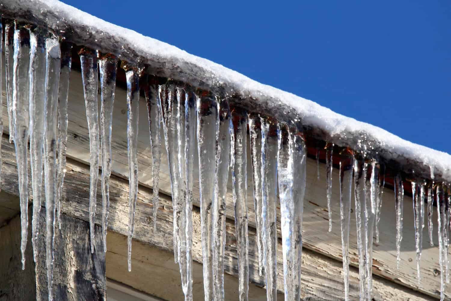 Icicles hanging from the edge of a snowy roof under a clear blue sky, illustrating winter weather challenges for Cape Cod homes.