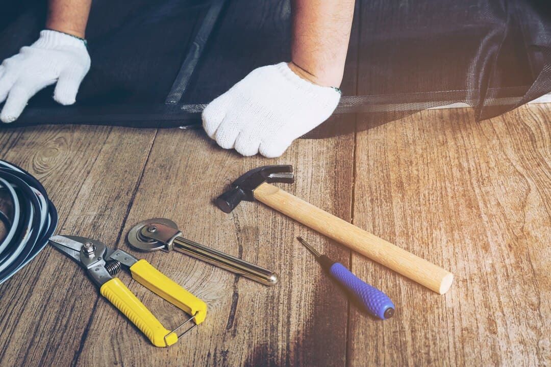 Carpenter's hands in white gloves working with tools including pliers, a hammer, and a screwdriver on a wooden surface, illustrating deck repair and maintenance.