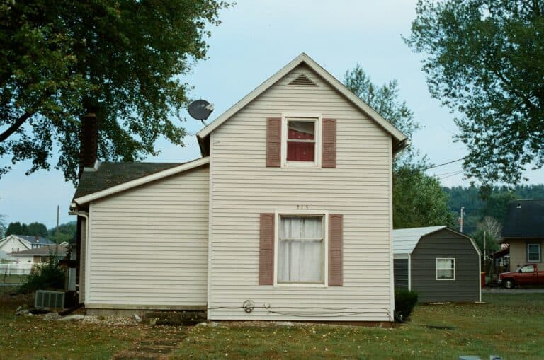 House with light-colored siding and red shutters, surrounded by trees, illustrating the impact of Cape Cod winters on home exteriors.