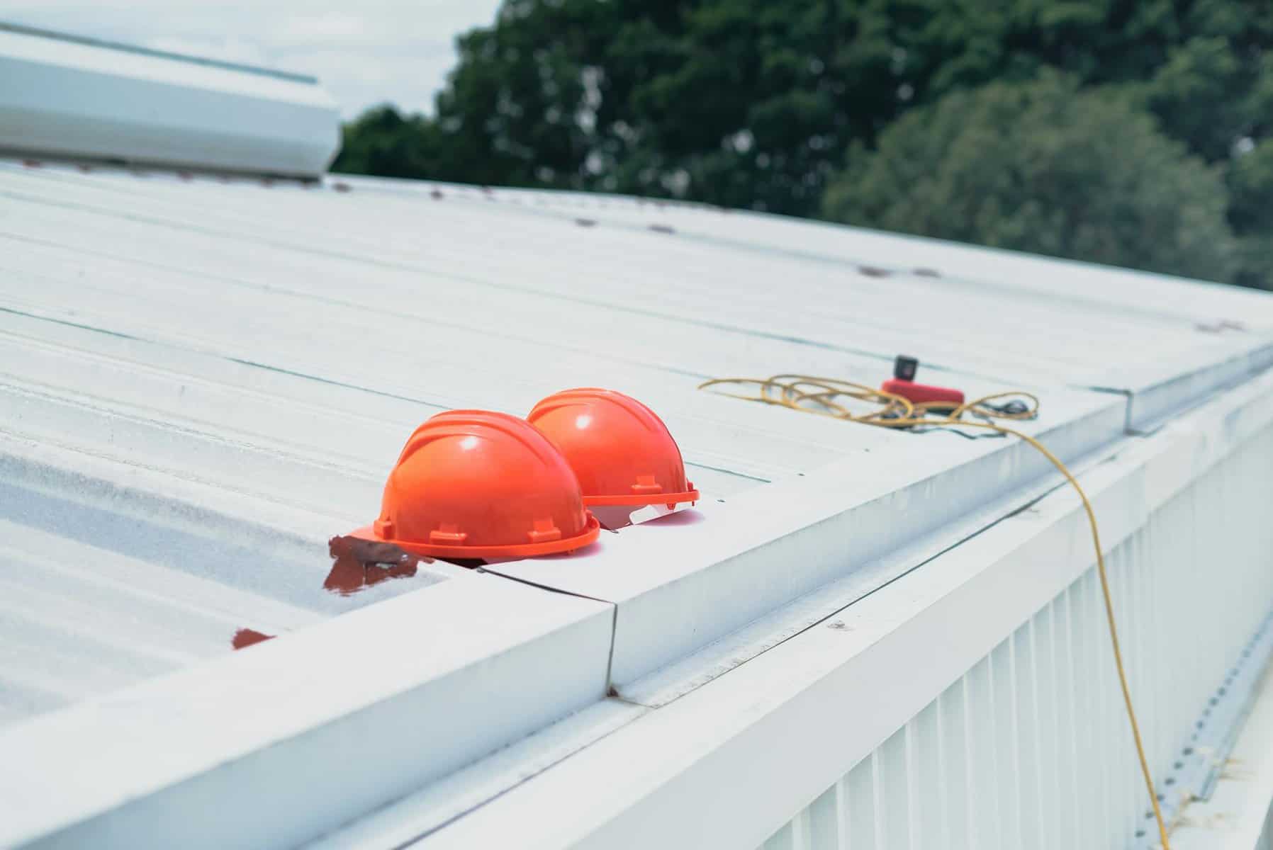 Two orange hard hats on a metal roof with a measuring tape and electrical cord, illustrating safety measures in roofing construction for Cape Cod projects.