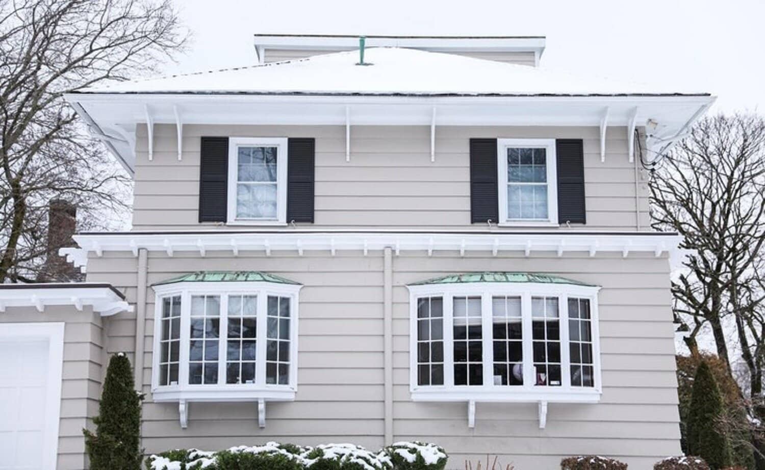 Snow-covered house with beige siding, black shutters, and bay windows, highlighting the importance of siding maintenance before winter in Cape Cod.